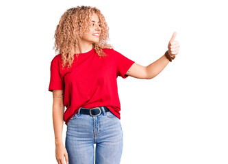 Young blonde woman with curly hair wearing casual red tshirt looking proud, smiling doing thumbs up gesture to the side