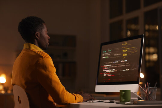 Side view portrait of contemporary African-American man looking at computer screen while working late at night writing code, copy space