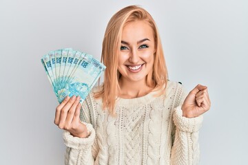 Beautiful caucasian blonde woman holding 100 brazilian real banknotes screaming proud, celebrating victory and success very excited with raised arm