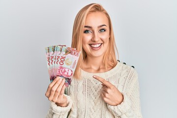 Beautiful caucasian woman holding 100 new zealand dollars banknote smiling happy pointing with hand and finger