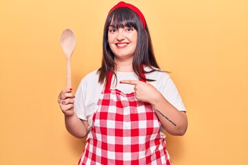 Young plus size woman wearing apron holding wooden spoon smiling happy pointing with hand and finger