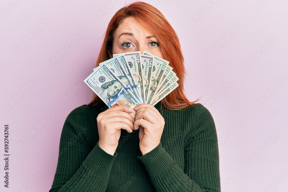 Poster Beautiful redhead woman holding dollars close to face skeptic and nervous, frowning upset because of problem. negative person.