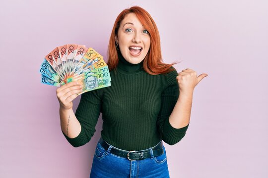 Beautiful redhead woman holding australian dollars pointing thumb up to the side smiling happy with open mouth