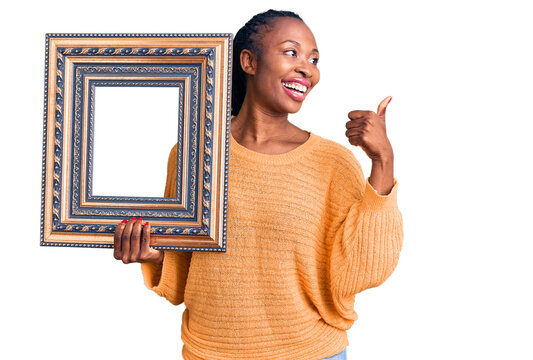 Young african american woman holding empty frame pointing thumb up to the side smiling happy with open mouth