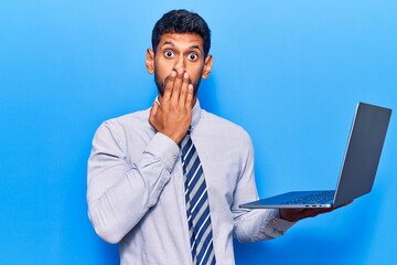 Young latin man holding laptop covering mouth with hand, shocked and afraid for mistake. surprised expression