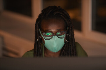 High angle portrait of contemporary African-American woman wearing mask while using computer lit by screen in dark office, copy space