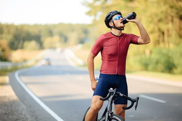 Tuinposter Muscular cyclist relaxing on bike and drinking water © Tymoshchuk