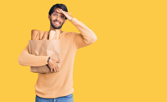 Handsome hispanic man holding paper bag with bread stressed and frustrated with hand on head, surprised and angry face