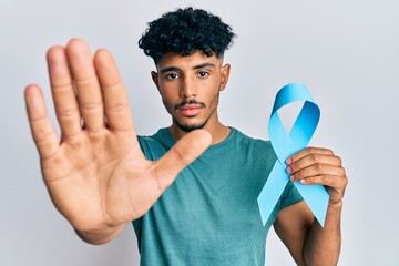 Young arab handsome man holding blue ribbon with open hand doing stop sign with serious and...