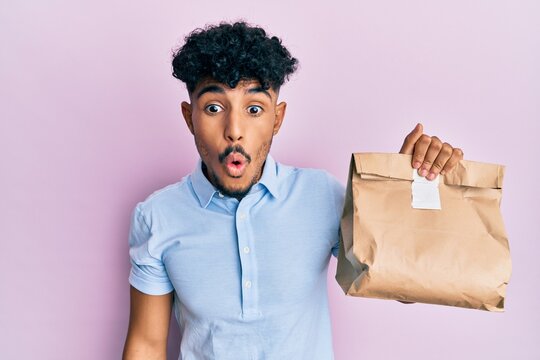 Young arab handsome man holding take away paper bag scared and amazed with open mouth for surprise, disbelief face