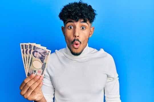 Young arab handsome man holding 5000 japanese yen banknotes scared and amazed with open mouth for surprise, disbelief face