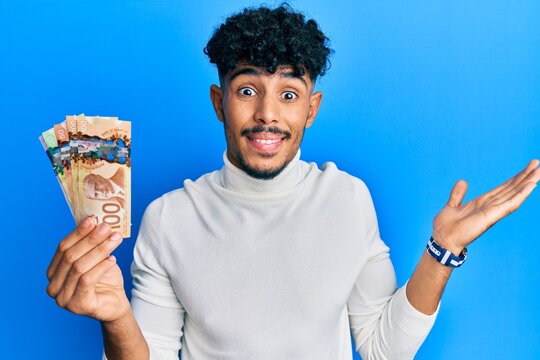 Young arab handsome man holding canadian dollars celebrating achievement with happy smile and winner expression with raised hand