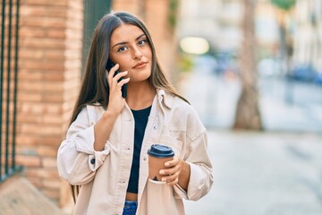 Young hispanic girl with serious expression talking on the smartphone and drinking take away coffee at the city.