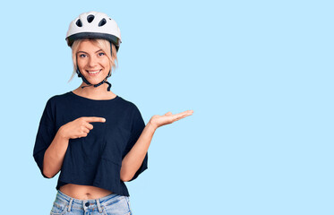Young beautiful blonde woman wearing bike helmet amazed and smiling to the camera while presenting with hand and pointing with finger.