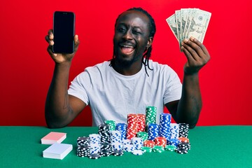 Handsome young black man playing poker holding smartphone and dollars winking looking at the camera...