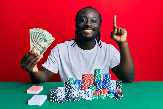 Handsome young black man playing poker holding 20 dollars banknotes smiling with an idea or question pointing finger with happy face, number one