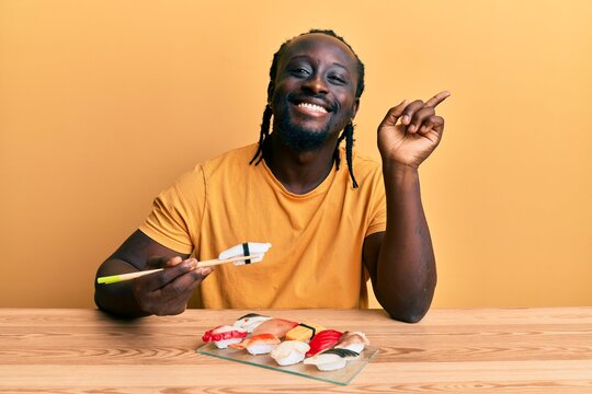 Handsome young black man eating sushi sitting on the table smiling happy pointing with hand and finger to the side