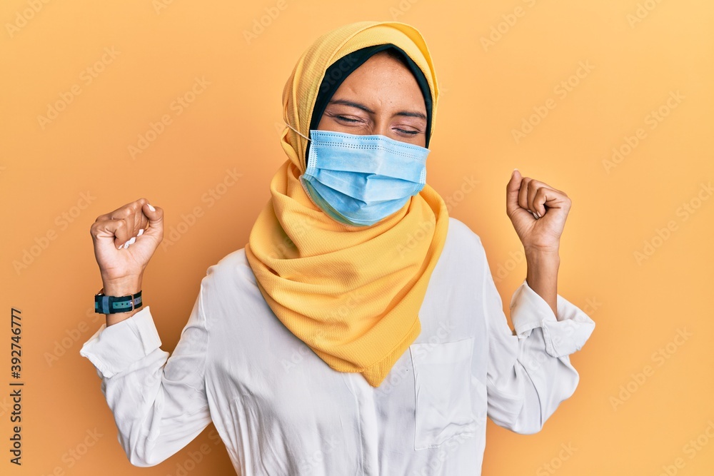 Poster Young brunette arab woman wearing traditional islamic hijab and medical mask very happy and excited doing winner gesture with arms raised, smiling and screaming for success. celebration concept.