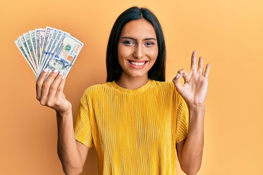 Young brunette woman holding dollars doing ok sign with fingers, smiling friendly gesturing excellent symbol