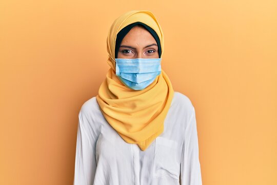 Young brunette arab woman wearing traditional islamic hijab and medical mask looking positive and happy standing and smiling with a confident smile showing teeth