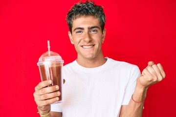 Young handsome man holding glass of smoothie screaming proud, celebrating victory and success very excited with raised arm