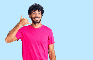 Handsome young man with curly hair and bear wearing casual pink tshirt smiling doing phone gesture with hand and fingers like talking on the telephone. communicating concepts.