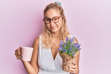 Young blonde girl holding cup of tea and herbal plant winking looking at the camera with sexy...
