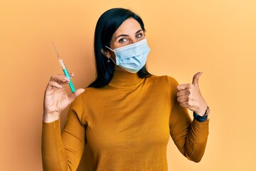 Young caucasian woman wearing medical mask holding syringe smiling happy and positive, thumb up doing excellent and approval sign