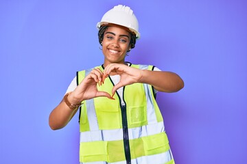 Young african american woman with braids wearing safety helmet and reflective jacket smiling in love showing heart symbol and shape with hands. romantic concept.