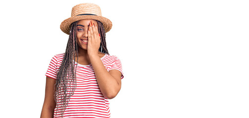 Obraz na płótnie Canvas Young african american woman with braids wearing summer hat covering one eye with hand, confident smile on face and surprise emotion.