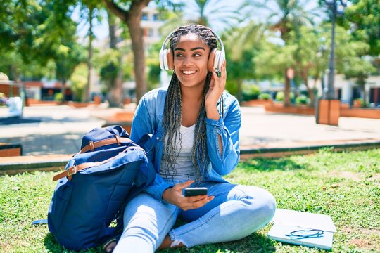 Young african american student woman smiling happy listening to music sitting on the grass at the university campus
