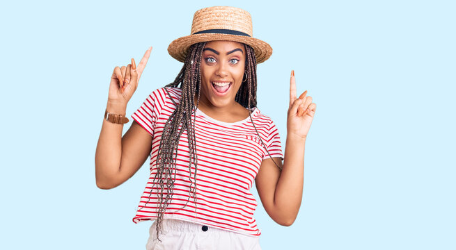 Young african american woman with braids wearing summer hat smiling amazed and surprised and pointing up with fingers and raised arms.