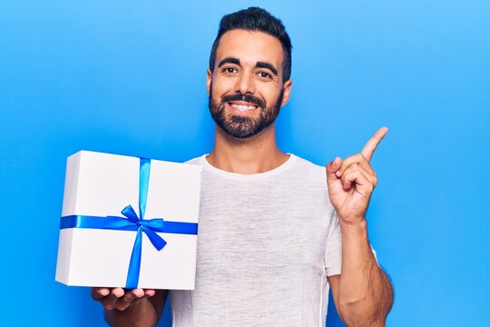 Young hispanic man holding gift smiling happy pointing with hand and finger to the side