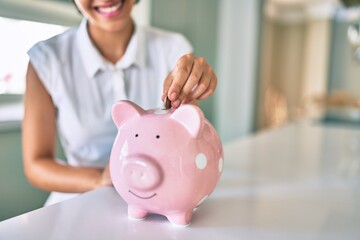 Young brunette woman smiling happy putting money savings inside of piggy bank