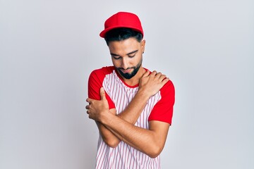 Obraz premium Young man with beard wearing baseball uniform hugging oneself happy and positive, smiling confident. self love and self care