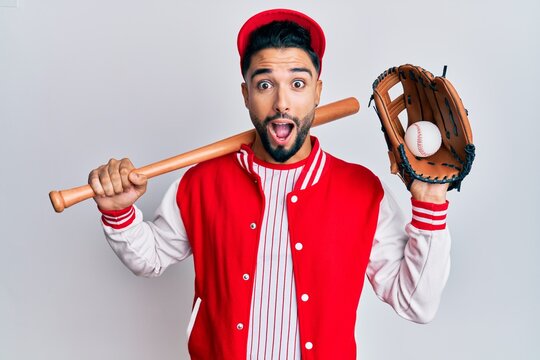 Young man with beard playing baseball holding bat and ball afraid and shocked with surprise and amazed expression, fear and excited face.