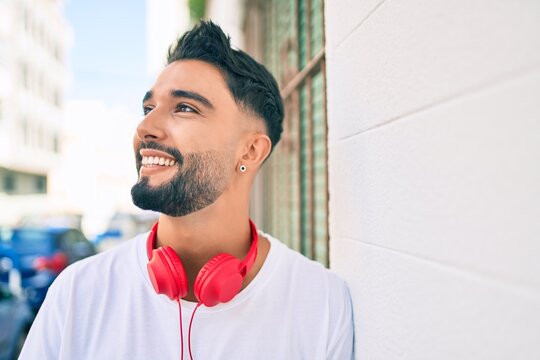 Young arab man smiling happy using headphones leaning on the wall at the city.