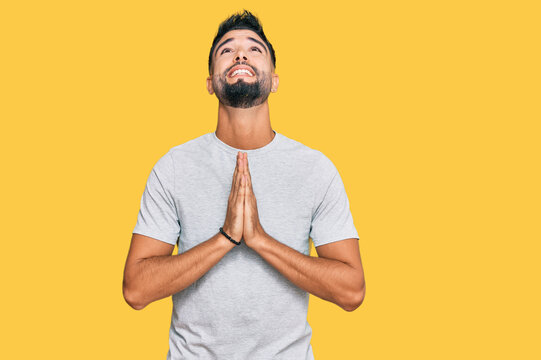 Young man with beard wearing casual grey tshirt begging and praying with hands together with hope expression on face very emotional and worried. begging.