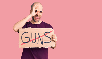 Young handsome man holding prohibited guns banner smiling happy doing ok sign with hand on eye looking through fingers