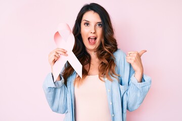 Young beautiful brunette woman holding pink cancer ribbon pointing thumb up to the side smiling happy with open mouth