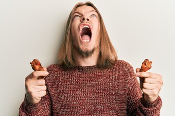 Handsome caucasian man with long hair eating chicken wings angry and mad screaming frustrated and...