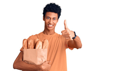 Young african american man holding paper bag with bread approving doing positive gesture with hand,...