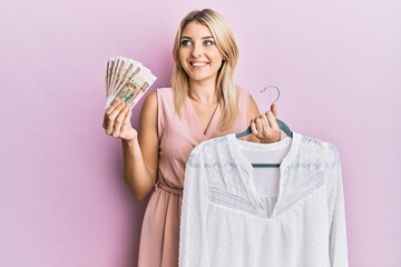 Young caucasian woman holding hanger with t shirt and russian ruble banknotes smiling looking to the side and staring away thinking.