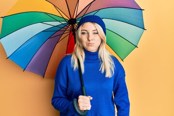 Young caucasian woman holding colorful umbrella puffing cheeks with funny face. mouth inflated with air, catching air.