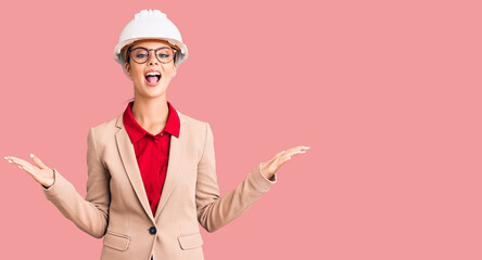 Young beautiful woman wearing architect hardhat celebrating victory with happy smile and winner expression with raised hands