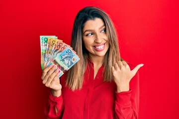 Beautiful brunette woman holding australian dollars smiling with happy face looking and pointing to the side with thumb up.
