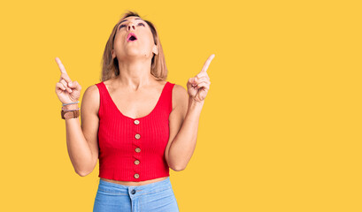 Young blonde woman wearing casual clothes amazed and surprised looking up and pointing with fingers and raised arms.