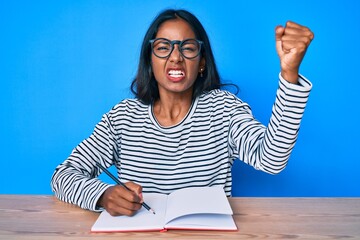 Young indian girl writing notebook sitting on the table annoyed and frustrated shouting with anger,...