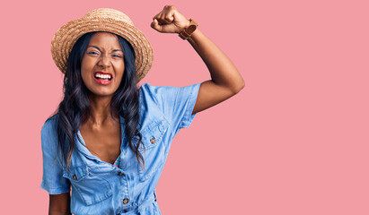 Young indian girl wearing summer hat angry and mad raising fist frustrated and furious while...