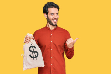 Young hispanic man holding dollars bag pointing thumb up to the side smiling happy with open mouth